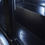 Interior of a dark metal cabinet or rack with smooth panels and a small circular access hole on the floor; visible hinges on the lower left.”,